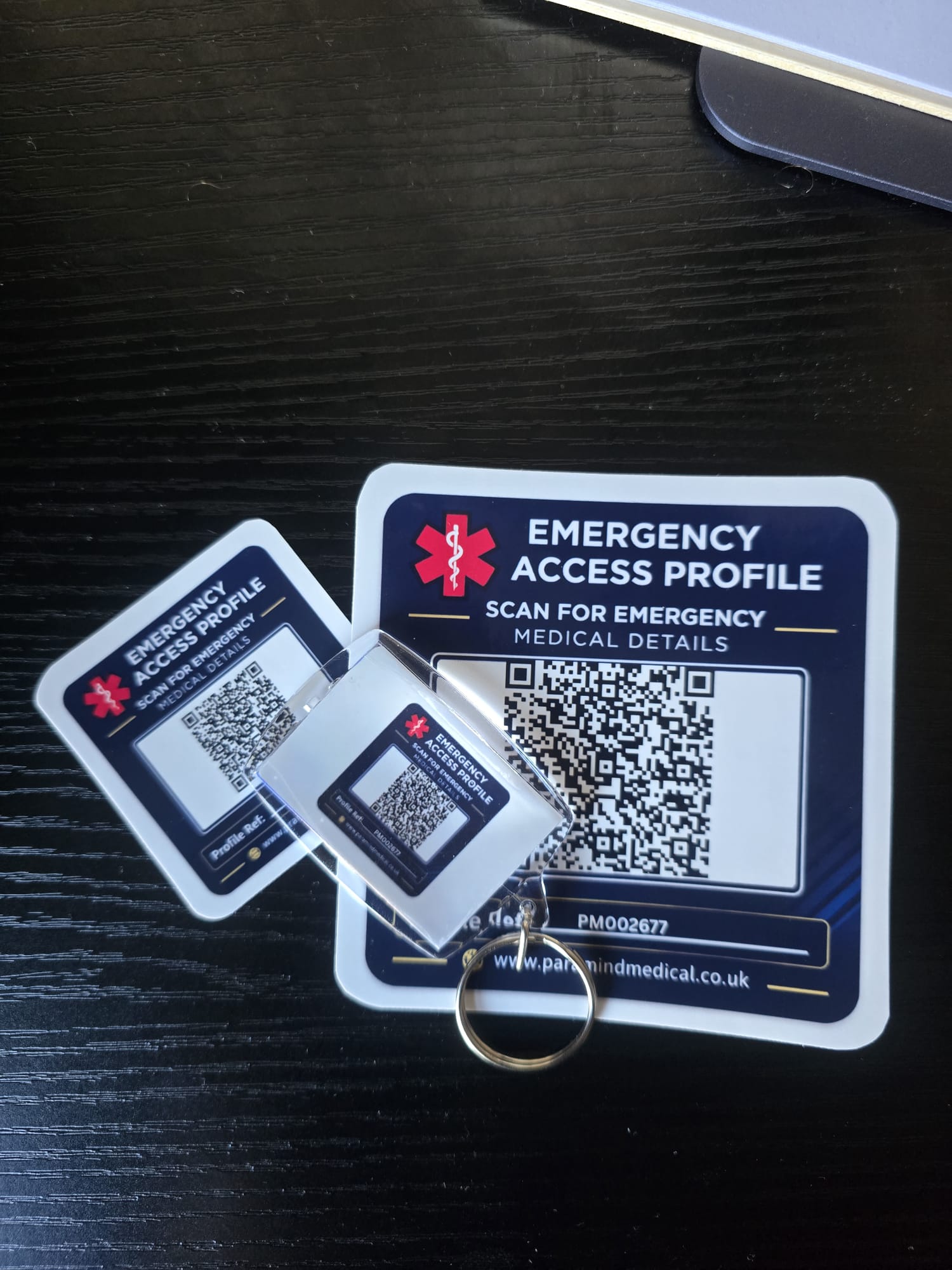 Paramind Medical Lifeline physical products: large emergency access profile QR sticker, small matching QR sticker, and clear acrylic keyring with Star of Life branding for UK emergency medical information
